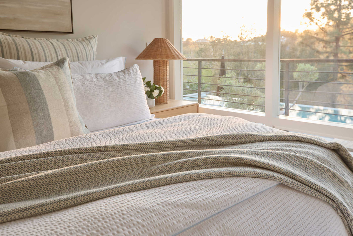 Blue-grey woven throw blanket with fringe, folded across bed linens