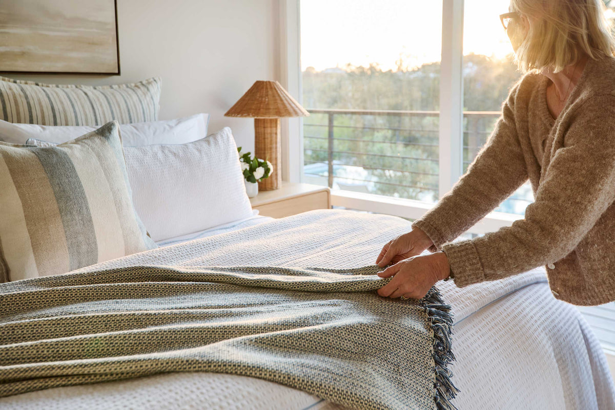 Blue-grey woven throw blanket with long fringe, laid across a bed
